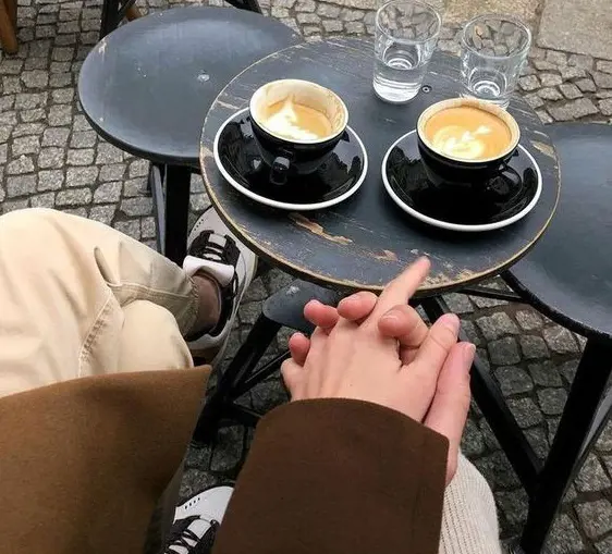 A couple holds hands while at a coffee shop where two cups of coffee is seen in the background. 