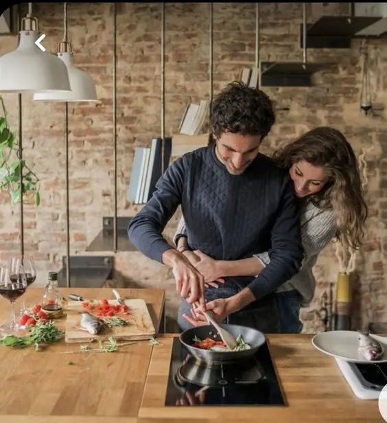 A young couple cooks together at a beautiful kitchen, boy stirs the pan while the girl is hugging her from behind. 