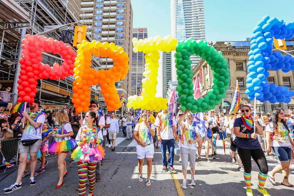 People participate in a Pride Parade with a sign reading Pride made with balloons representing the Rainbow Flag. 